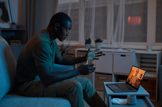 Hombre africano hablando con el terapeuta en línea en la computadora portátil en el cuarto oscuro en casa