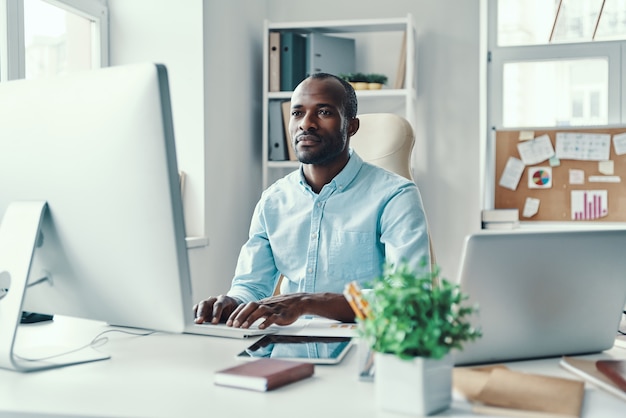 Hombre africano joven concentrado en camisa usando la computadora mientras trabajaba en la oficina