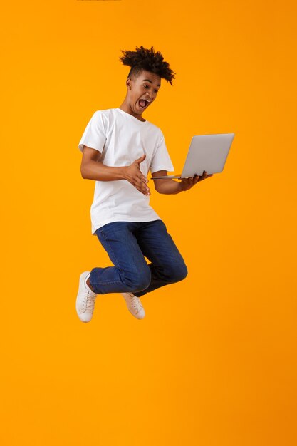 Hombre africano joven emocionado aislado sobre el espacio amarillo usando la computadora portátil.