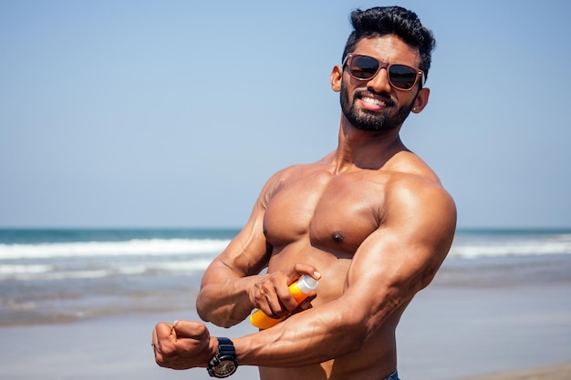 Hombre africano joven feliz en la playa. Culturista de fitness masculino guapo y confiado con sixpack. Modelo de India masculino perfecto abs, hombros, bíceps, tríceps, pecho sosteniendo una botella de protector solar