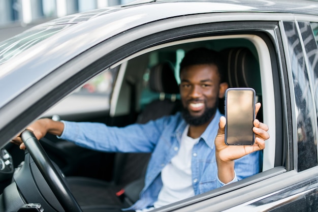 Hombre africano joven que sostiene el teléfono con la pantalla mientras está sentado en su coche. Copie el espacio.