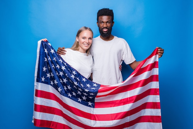 Hombre africano con mujer caucásica sosteniendo la bandera americana aislada en la pared azul