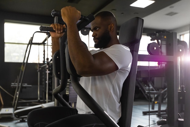 Un hombre africano musculoso y en forma entrena los músculos pectorales en un simulador de bloques en el gimnasio