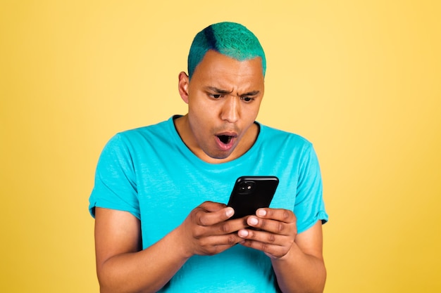 Hombre africano negro en casual en la pared amarilla con mirada de teléfono móvil en la pantalla sorprendido con la boca abierta