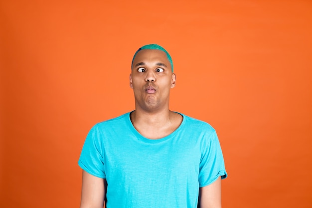 Hombre africano negro en casual en la pared naranja cabello azul hace muecas locas divertidas cara de pez divirtiéndose