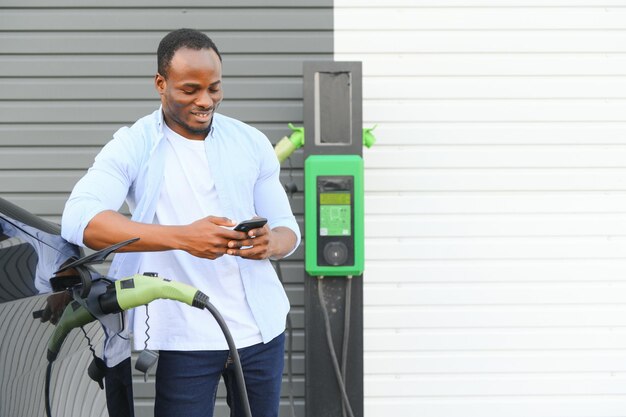 Hombre africano parado cerca de un coche eléctrico con cable de carga en el enchufe Vehículo ecológico cargando en el concepto de estilos de vida de la estación