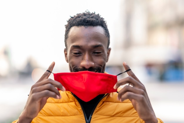 Foto hombre africano poniéndose una mascarilla roja con fondo de ciudad desenfocada