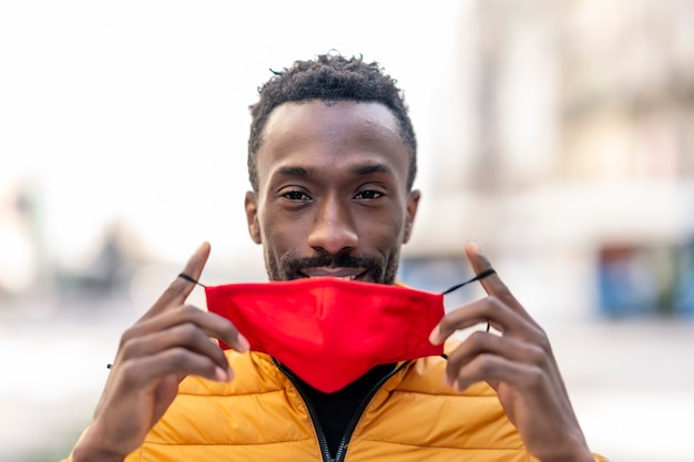 Foto hombre africano poniéndose una mascarilla roja con fondo de ciudad desenfocada