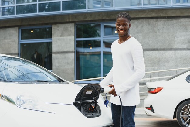 Hombre africano que sostiene el cable de carga en la mano de pie cerca de un coche eléctrico de lujo.