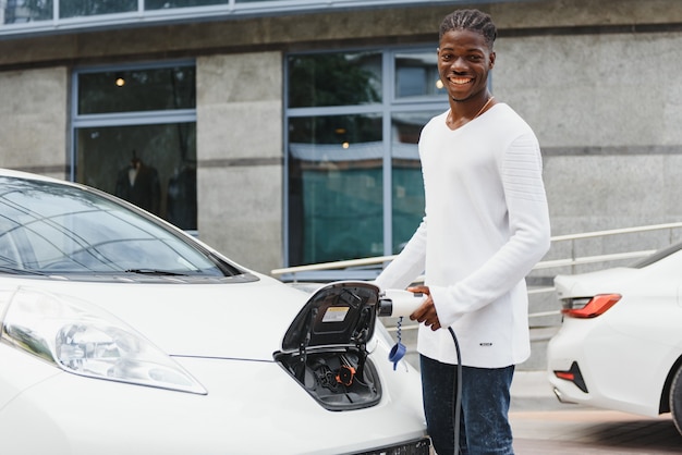 Hombre africano que sostiene el cable de carga en la mano de pie cerca de un coche eléctrico de lujo.