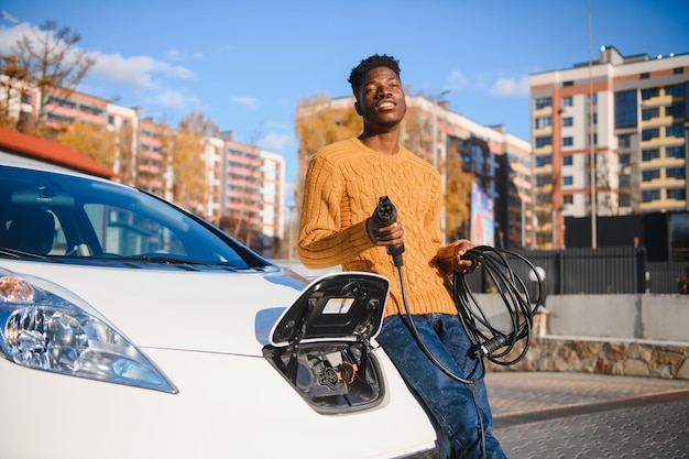 Hombre africano que sostiene el cable de carga en la mano de pie cerca de un coche eléctrico de lujo.