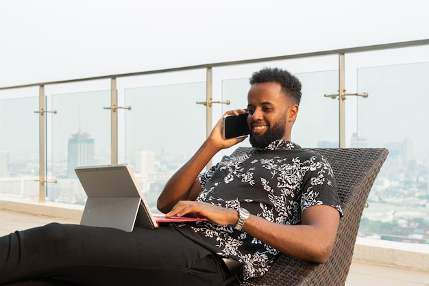 Hombre africano relajándose al aire libre mientras usa una computadora portátil y un teléfono móvil