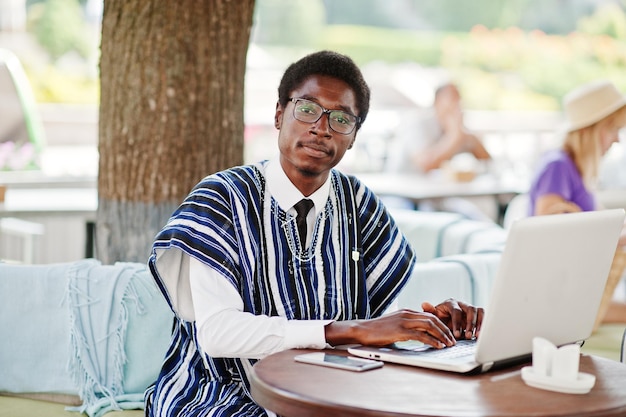 Hombre africano con ropa tradicional y anteojos sentado detrás de una laptop en un café al aire libre y trabajando