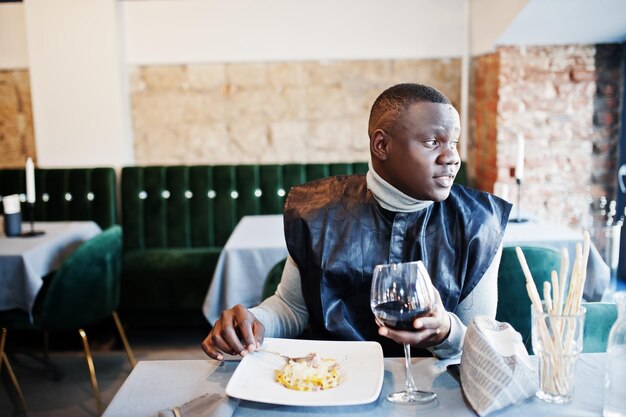 Hombre africano con ropa tradicional negra sentado en el restaurante y comiendo pasta y bebiendo vino