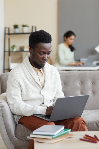 Hombre africano sentado en el sofá frente a la mesa con libros y escribiendo en la computadora portátil que está aprendiendo en línea en casa