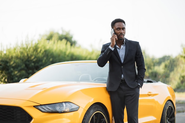 Hombre africano serio en traje sosteniendo un teléfono inteligente moderno en la mano mientras está de pie cerca de un auto descapotable