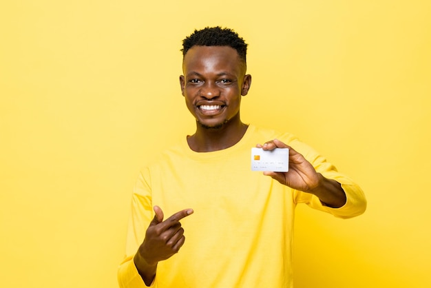 Hombre africano sonriente con atuendo informal apuntando a la tarjeta de crédito en un estudio aislado de color amarillo