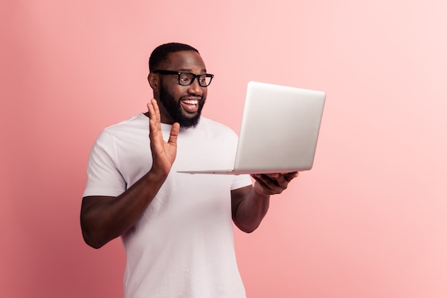 Hombre africano sonriente joven de pie y usando la computadora portátil hacer llamada mano de onda