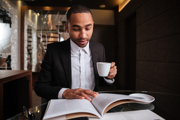 Hombre africano en traje sentado con diario y café en el hotel