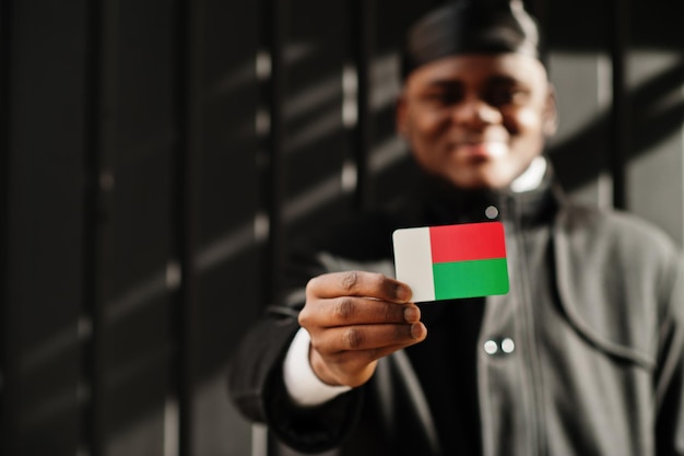 El hombre africano usa durag negro y sostiene la bandera de Madagascar en un fondo oscuro aislado a mano