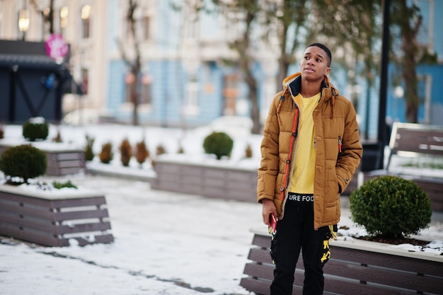 Hombre africano vestido con chaqueta naranja en el clima frío de invierno posado al aire libre con teléfono móvil