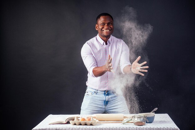 Hombre afro africano guapo y joven preparando pasteles caseros American Pie de manos de masa fresca sucias por harina, en la mesa hay huevos, rodillo y libro de recetas sobre un fondo negro en el estudio