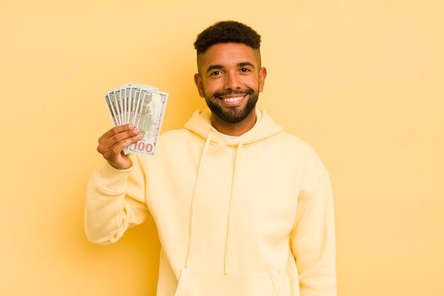 Hombre afro cool sonriendo felizmente con una mano en la cadera y un concepto de billete de dólar confiado