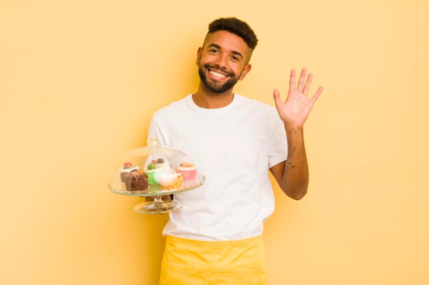 Hombre afro negro sonriendo felizmente saludando con la mano dándole la bienvenida y saludando el concepto de pasteles caseros