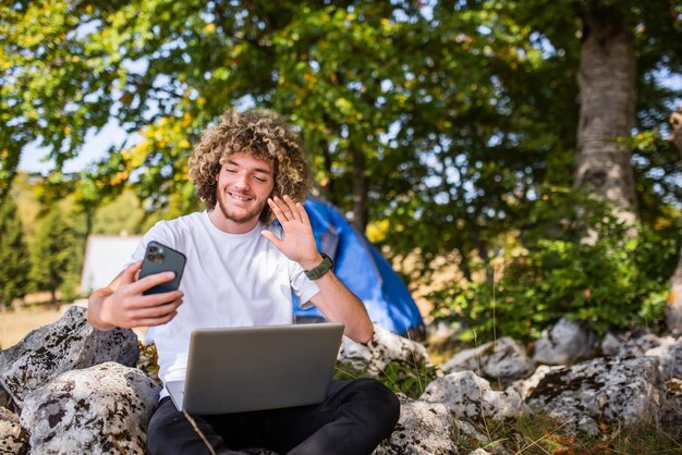 Un hombre afro se sienta en la naturaleza y usa un teléfono inteligente y una computadora portátil para una reunión en línea