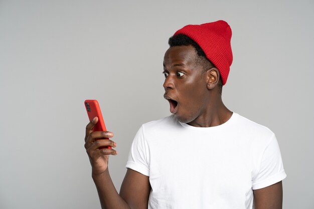 Foto hombre afro sorprendido hace grandes ojos abre la boca ampliamente, sorprendido con una foto indecente, mirando el móvil