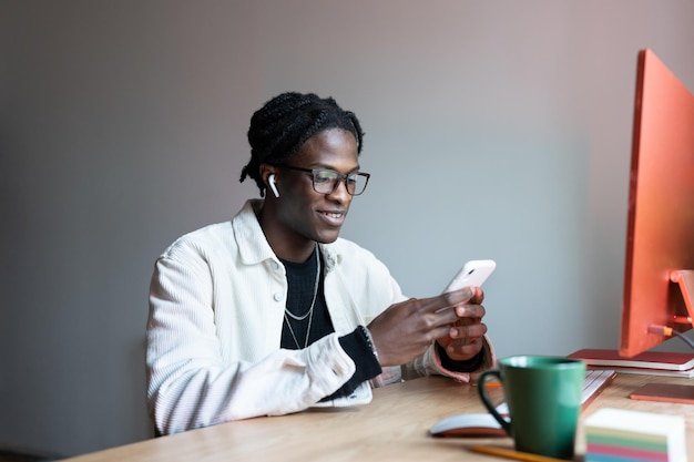 Un hombre afroamericano alegre y despreocupado con auriculares usa el teléfono para cambiar la lista de reproducción y se sienta en el escritorio de la oficina