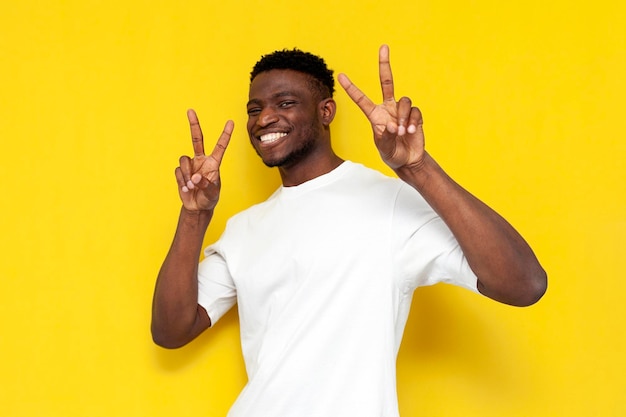 Hombre afroamericano alegre muestra gesto de paz con las manos y saluda sobre fondo amarillo aislado