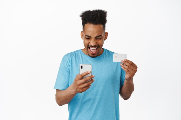 Hombre afroamericano alegre que mira el teléfono inteligente y la tarjeta de crédito, paga, compra en línea, sonríe emocionado, hace la compra, se enfrenta a un fondo blanco.