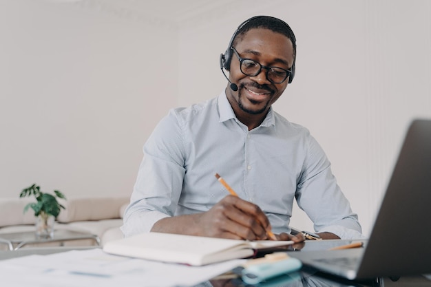 Hombre afroamericano en auriculares aprende en línea en la computadora portátil toma notas Educación a distancia de aprendizaje electrónico