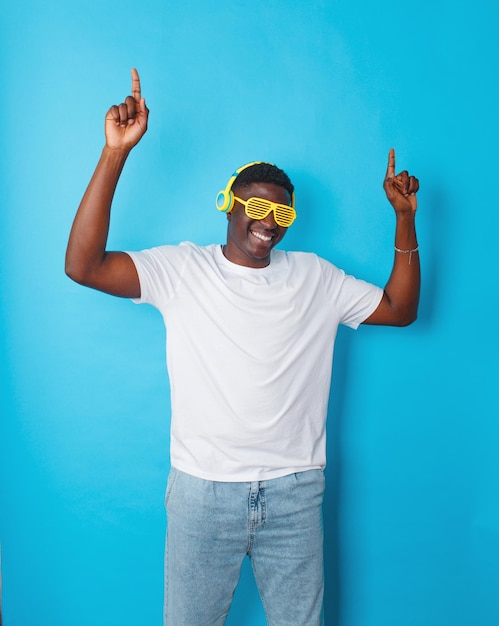 Un hombre afroamericano baila alegremente con auriculares amarillos y gafas elegantes en un fondo azul aislado