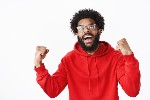 Hombre afroamericano con barba en sudadera con capucha roja levantando los puños en el éxito celebrando la victoria, triunfando de finalmente hacer realidad el sueño, mirando hacia arriba.