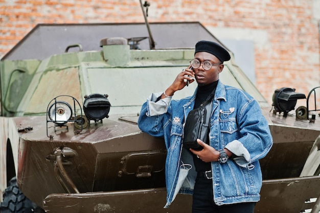 Hombre afroamericano con boina de chaqueta de jeans y anteojos hablando por teléfono contra el vehículo blindado militar btr