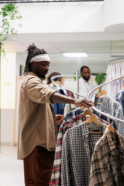 Hombre afroamericano en busca de ropa de moda en la tienda de ropa. Cliente navegando a través de camisas colgadas en el estante para elegir ropa informal en la boutique de moda del centro comercial