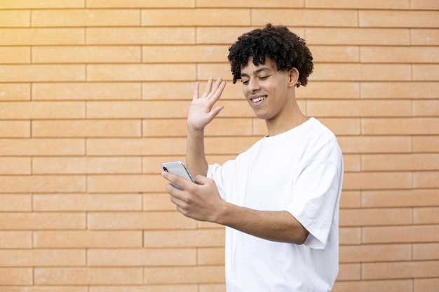 Hombre afroamericano de cabello rizado sonriente sosteniendo un teléfono y saludando a la cámara