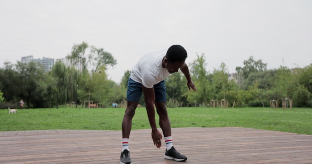 Hombre afroamericano calentando con ejercicios antes de un entrenamiento intenso al aire libre. Calentamiento de los músculos antes de trotar. Deportes al aire libre.