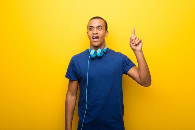 Hombre afroamericano con camiseta azul sobre fondo amarillo pensando en una idea apuntando el dedo hacia arriba