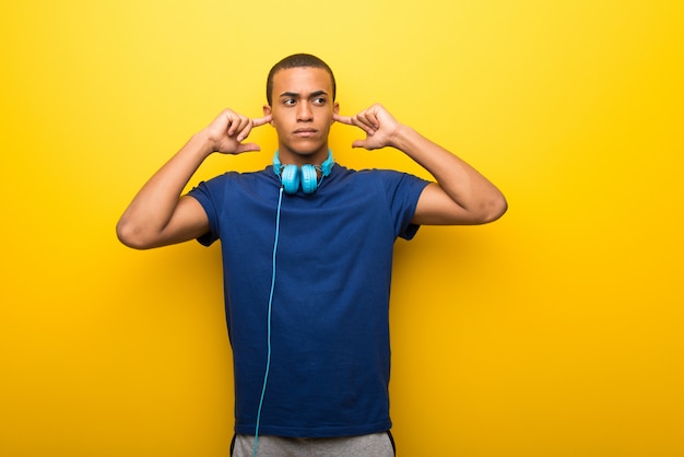 Hombre afroamericano con camiseta azul sobre fondo amarillo que cubre ambas orejas con las manos