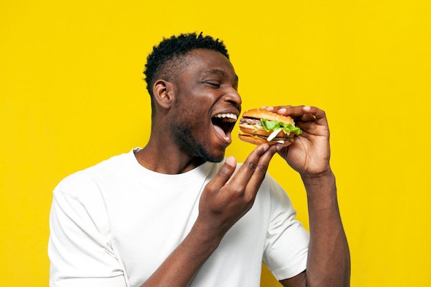 hombre afroamericano en camiseta blanca muerde una hamburguesa grande y se regocija en el fondo aislado amarillo