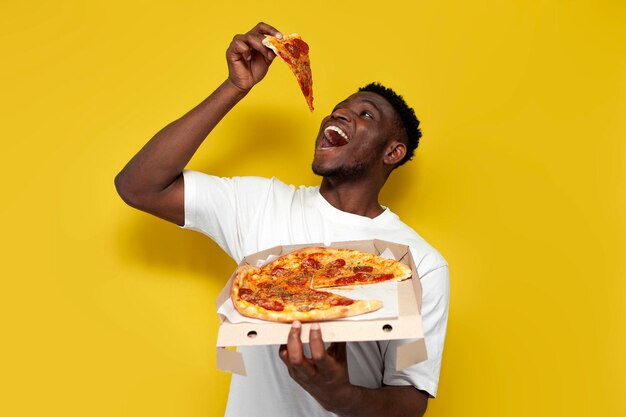 hombre afroamericano en camiseta blanca sostiene una caja de pizza y sonríe en fondo aislado amarillo