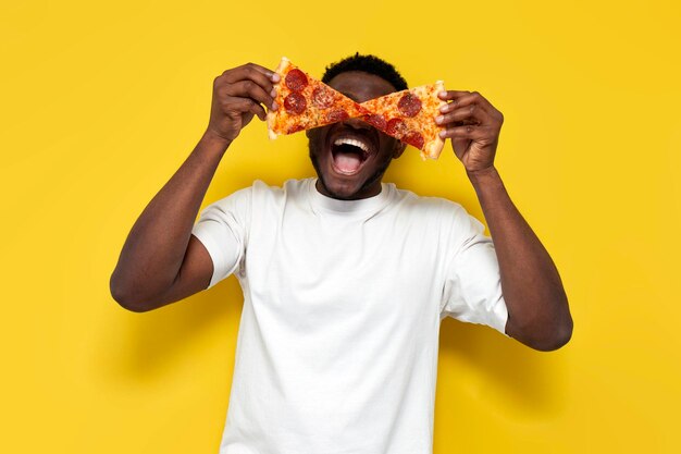 Hombre afroamericano en camiseta blanca sostiene dos pedazos de pizza delante de sus ojos y grita