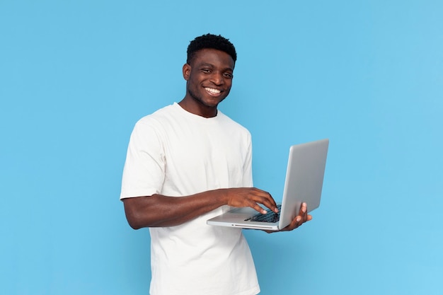 Un hombre afroamericano con camiseta blanca usa una computadora portátil y sonríe con un fondo azul aislado