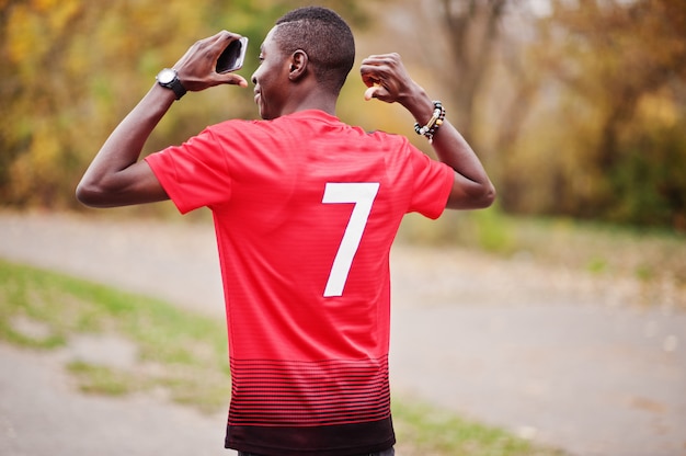 Hombre afroamericano en camiseta deportiva de fútbol rojo con número 7 contra el parque de otoño.