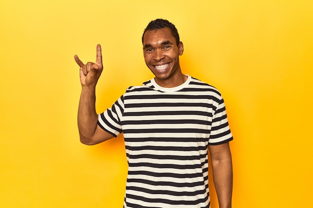 Hombre afroamericano en camiseta a rayas de estudio amarillo mostrando un gesto de cuernos como un concepto de revolución