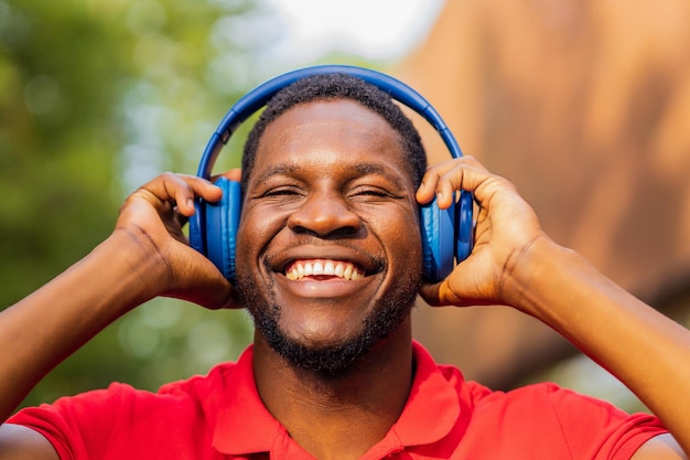 Hombre afroamericano en camiseta roja escuchando música en auriculares azules y estando de buen humor al aire libre