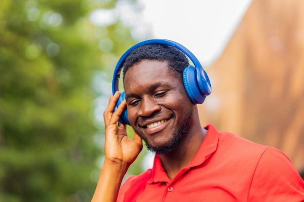 Hombre afroamericano en camiseta roja escuchando música en auriculares azules y estando de buen humor al aire libre